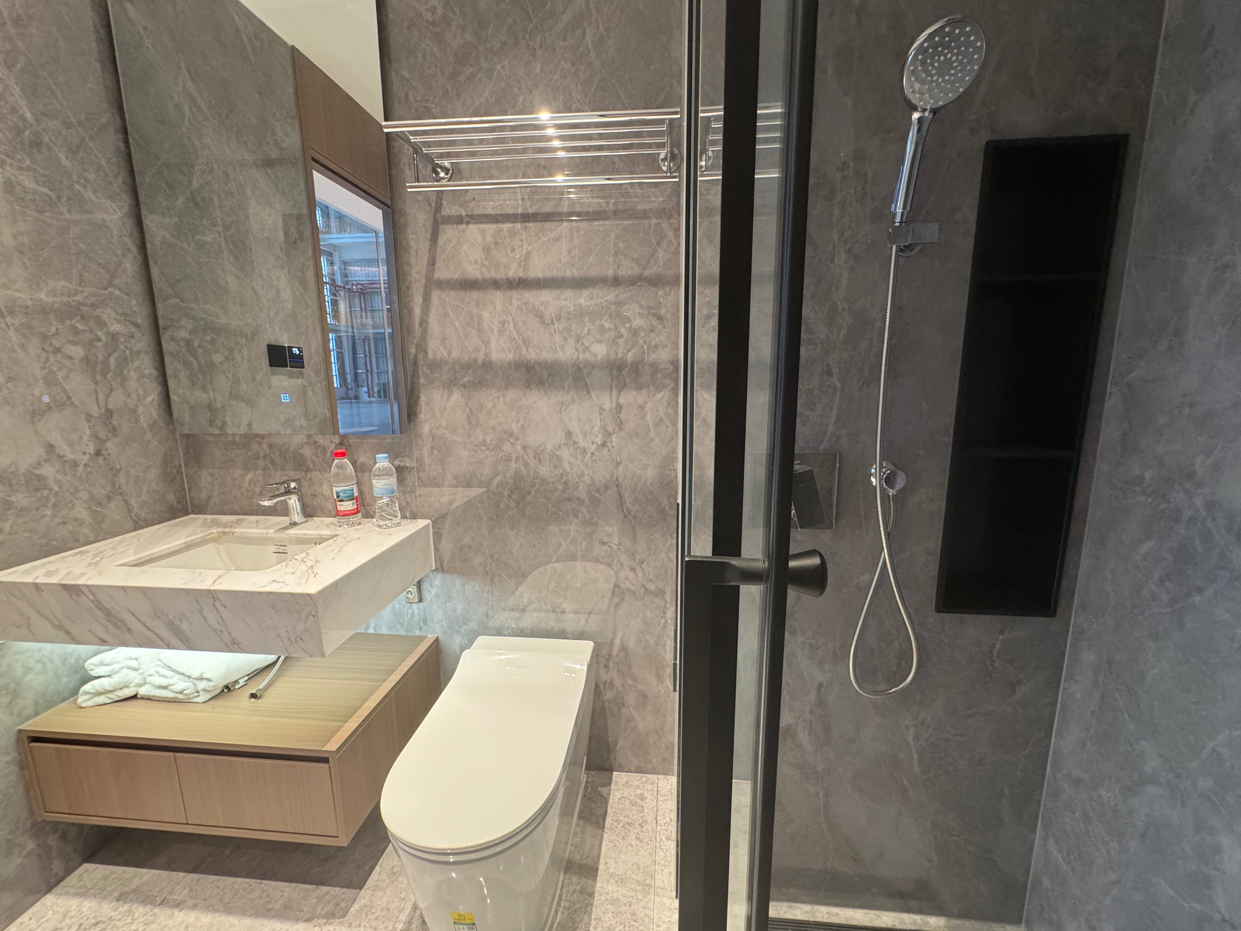 Modern modular hotel bathroom interior with elegant grey marble-patterned wall tiles, a floating stone sink vanity, a smart toilet, and a glass-enclosed shower stall with black metal frames and chrome fixtures.