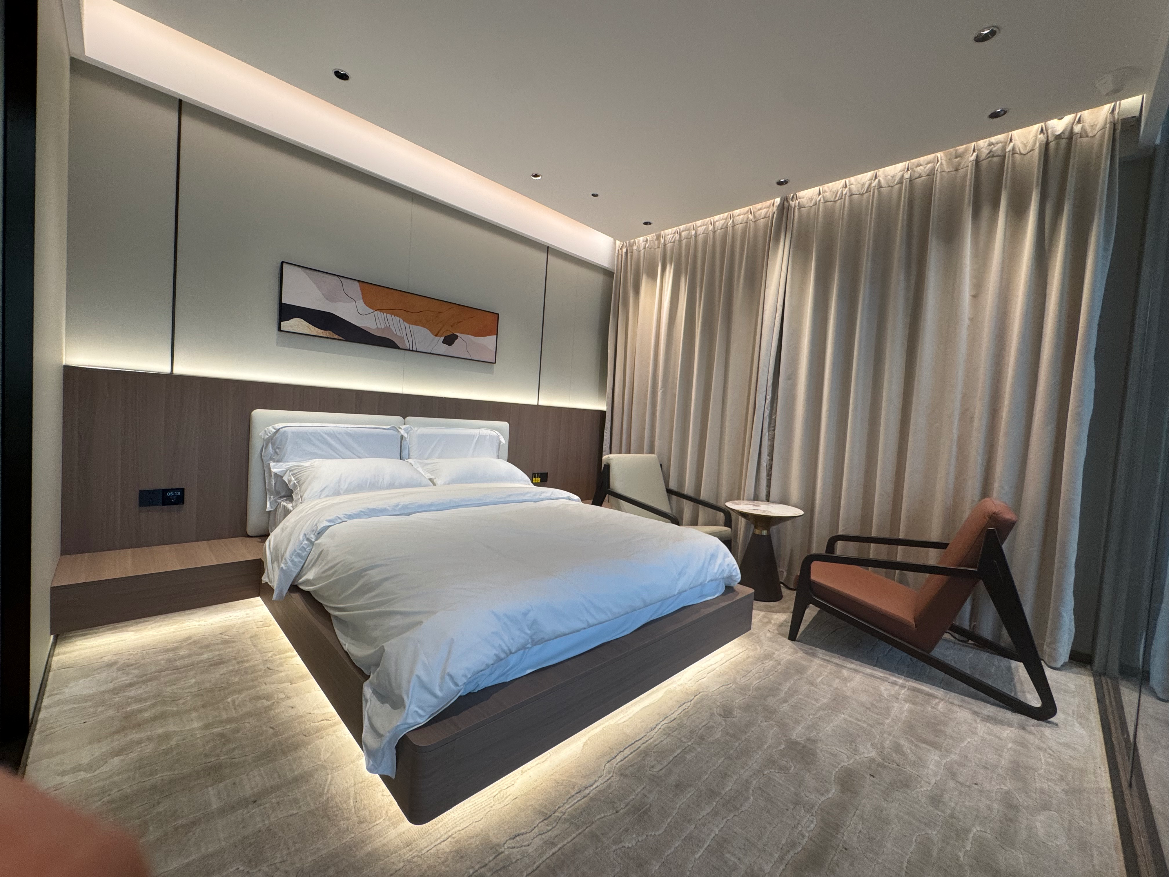 Interior of a modern modular hotel bedroom featuring a minimalist floating bed with warm under-bed LED lighting, a wooden accent headboard wall, abstract decor art, and a seating area with two contemporary armchairs.
