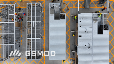 Overhead view of steel-framed modular building units at different stages of factory assembly, showing structural frames, metal decking and workers.