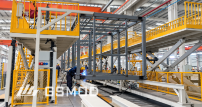 Workers welding a rectangular steel building module frame on an elevated production platform inside a manufacturing facility.